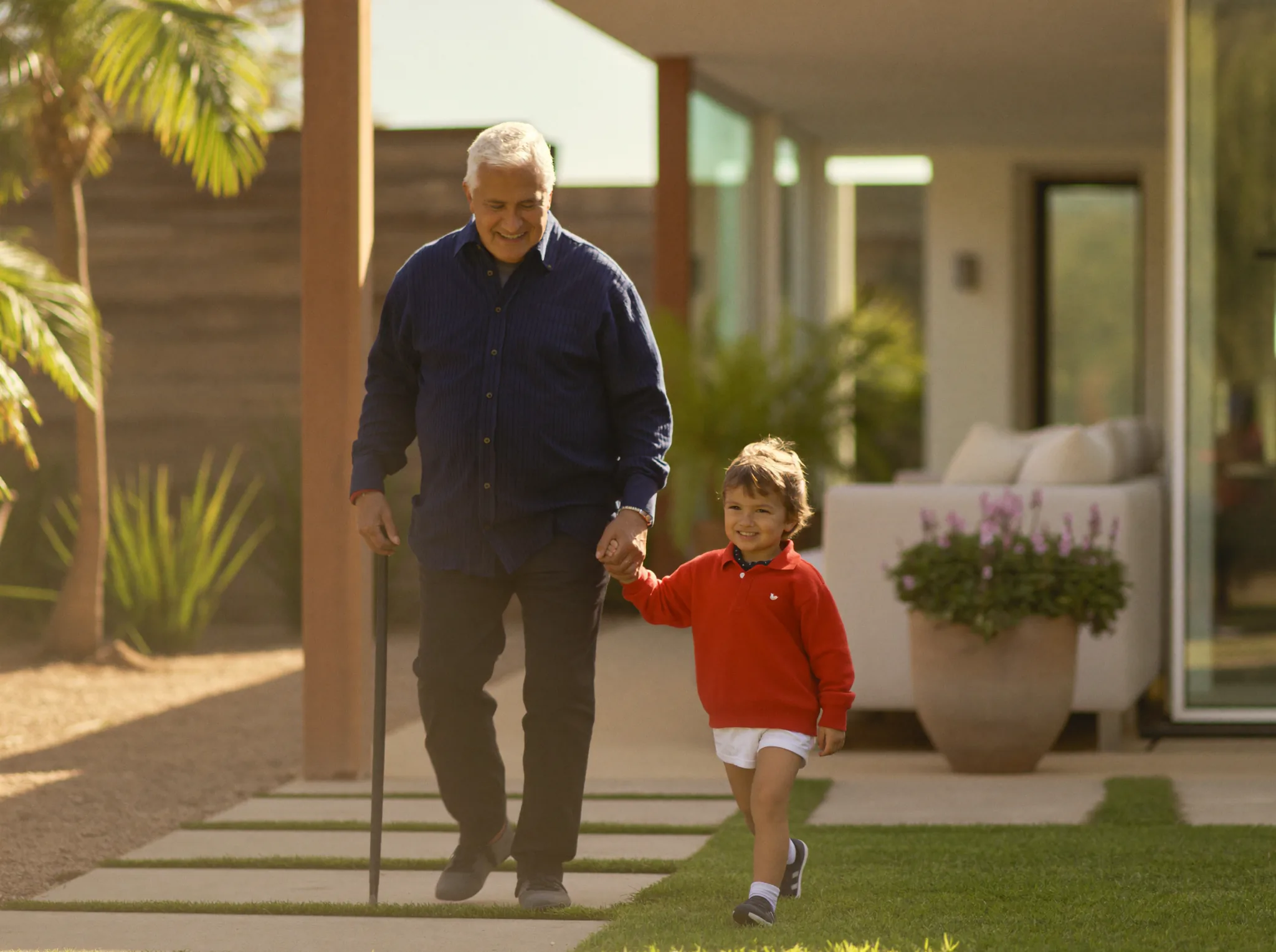 A senior man and his young grandson walking through a sunlit garden holding hands