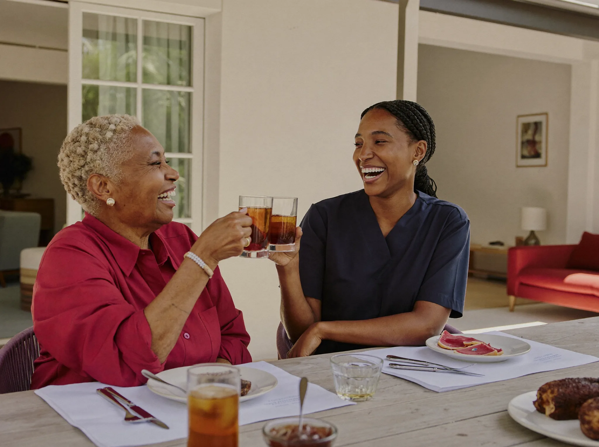 A senior and her caregiver laughing together over iced tea in a sunlit garden