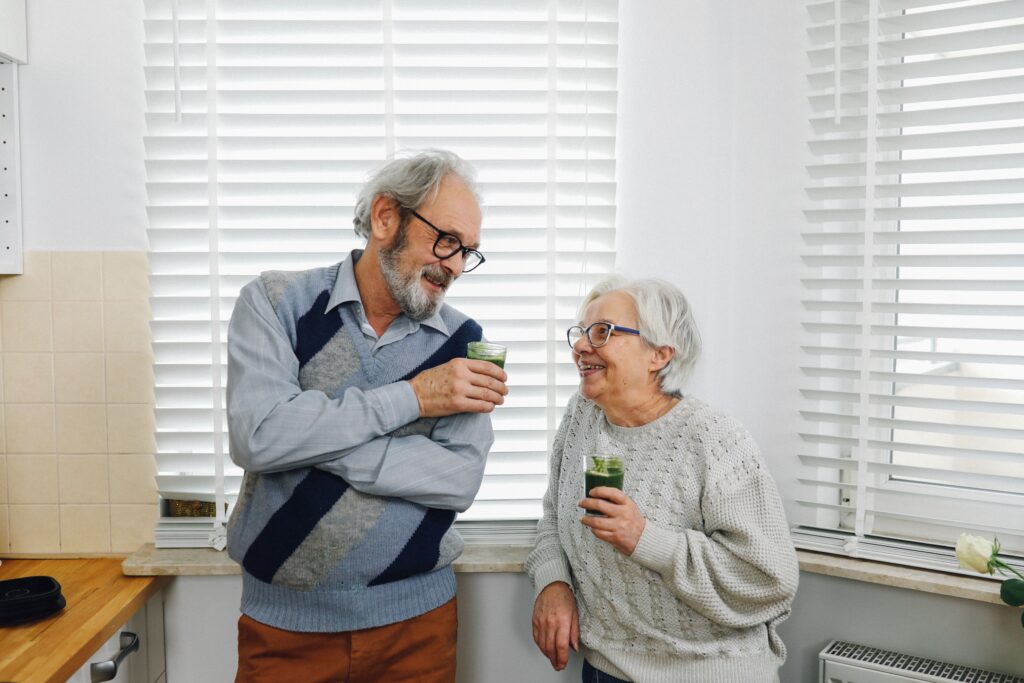 An older senior couple in a kitchen smiling and talking while holding glasses of green vegetable juice, representing healthy diet choices and gut health for seniors managing autoimmune gastritis.