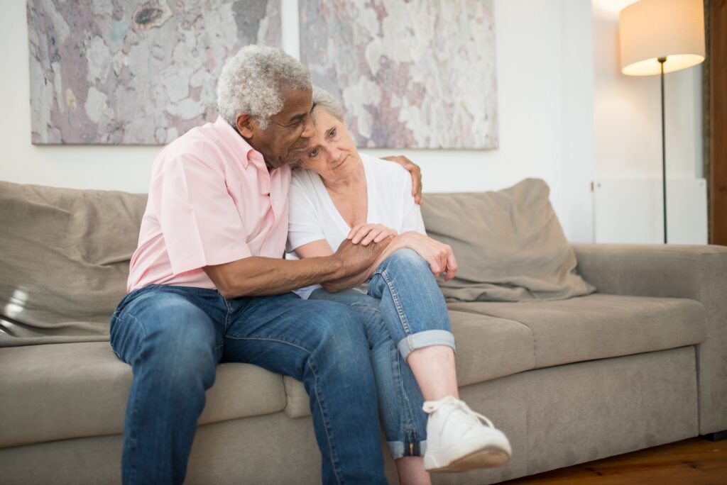 An older Black man comfortably comforting a concerned older woman on a couch, representing family support and health awareness for seniors in Alabama.